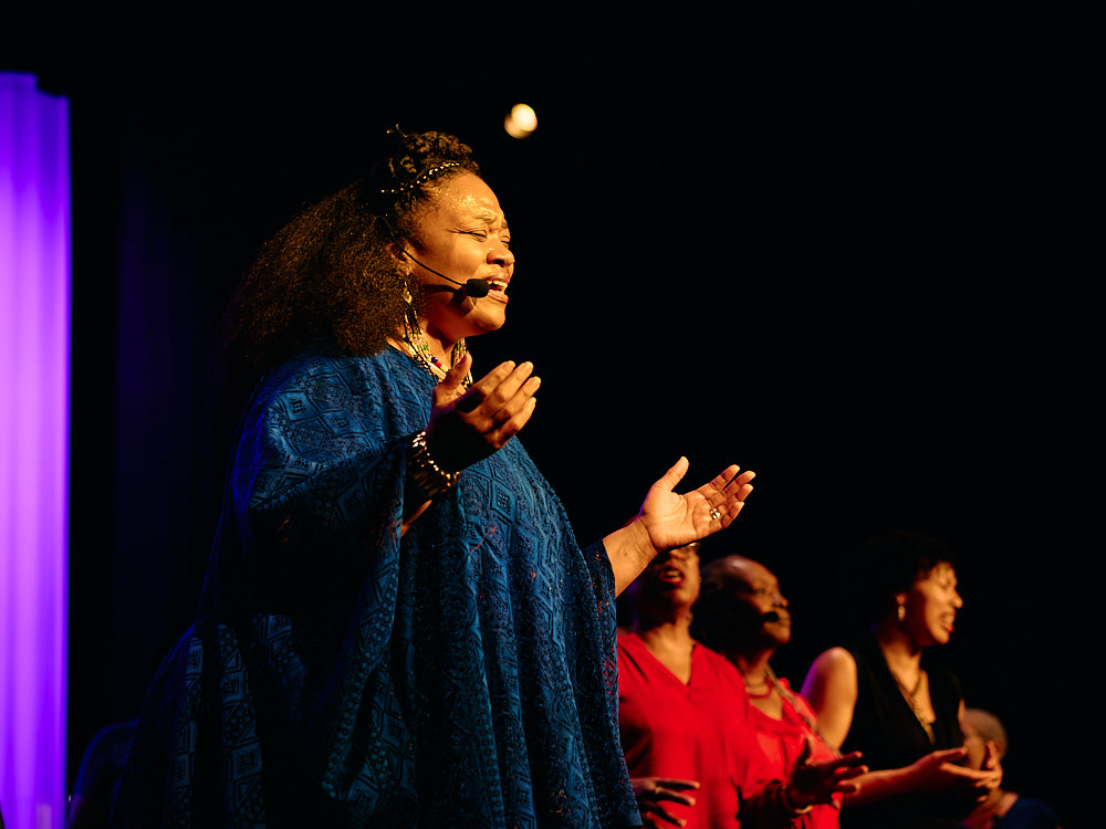 A host speaks on stage in front of a purple-lit curtain, addressing the audience.