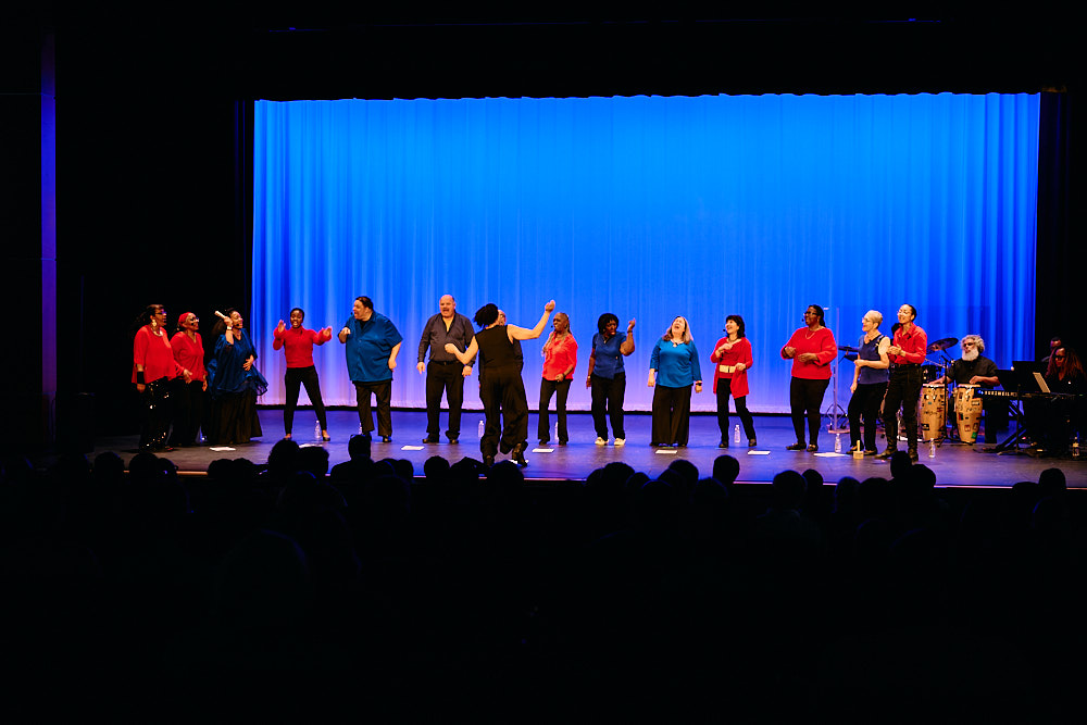 The full Exult Choir lined up on stage under blue lighting, with a musician on keyboard at the side.