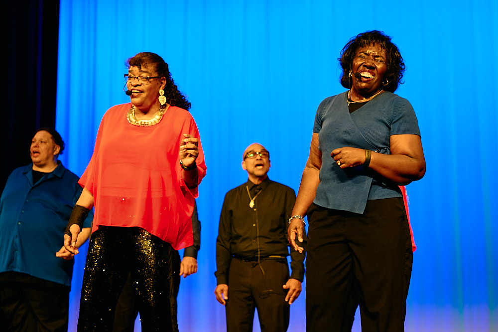 Two performers sing together on stage under blue lighting.